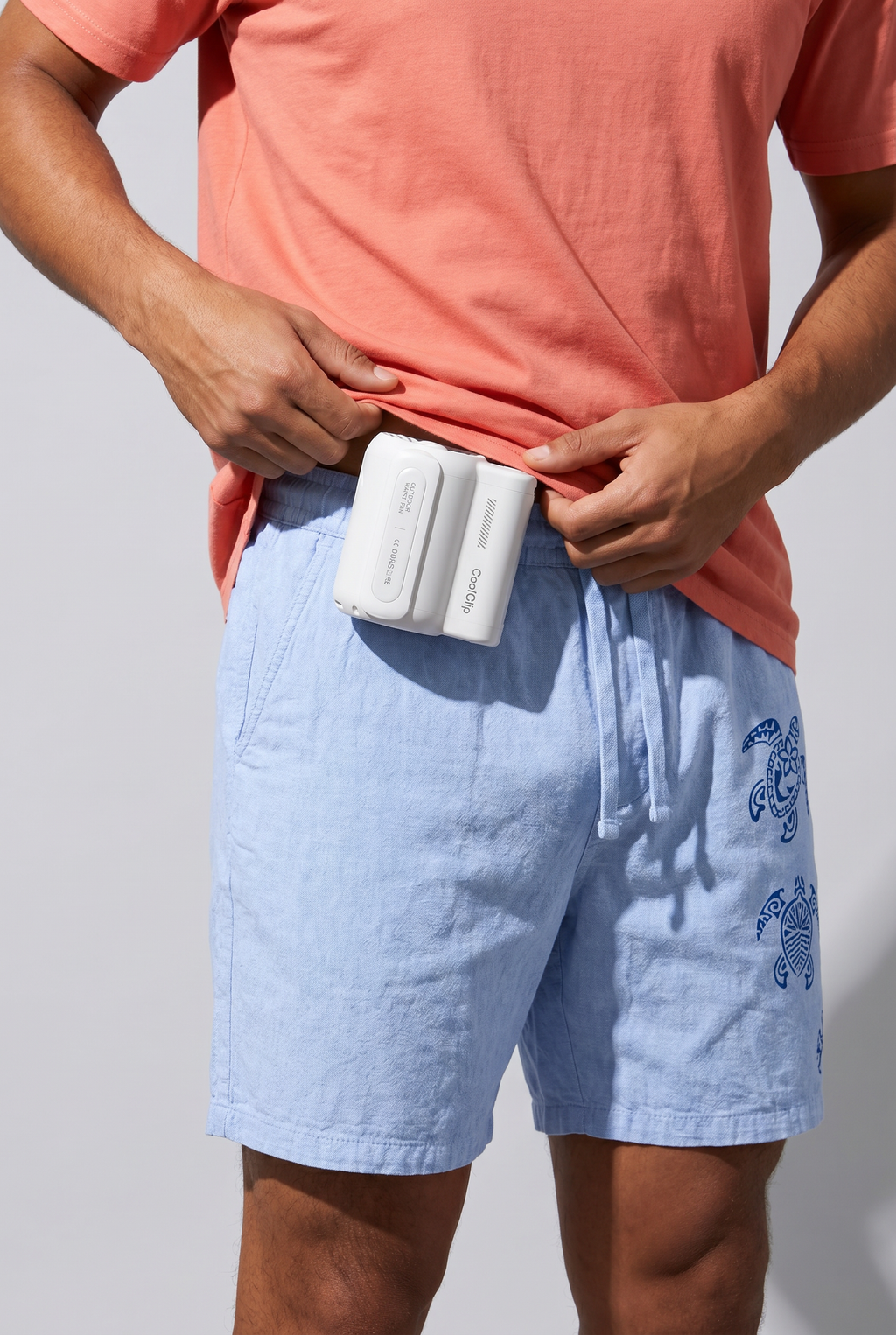 Person wearing light blue shorts with a white CoolClip portable fan on their waist against a gray background